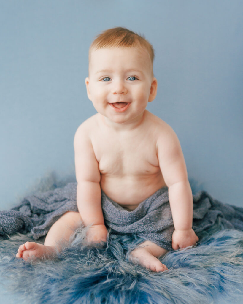 six month old baby boy smiling at camera sitting up on blue blankets and blue backdrop