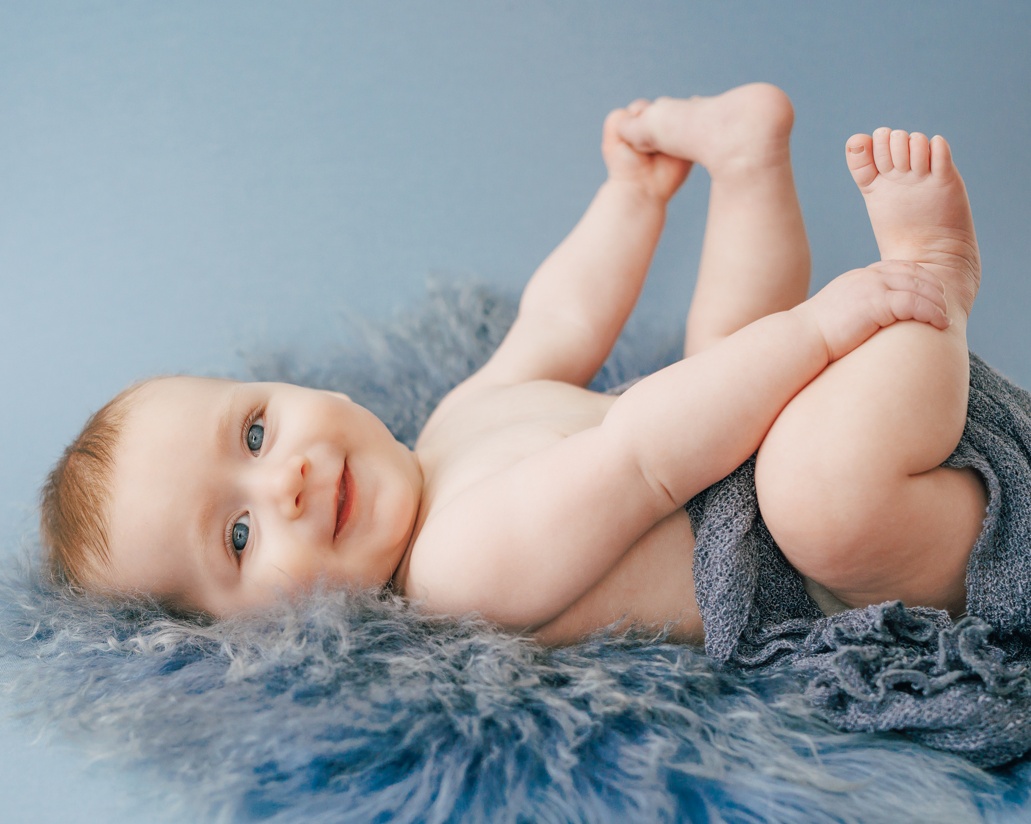 six month old baby boy swaddled in blue cloth holding feet on blue backdrop
