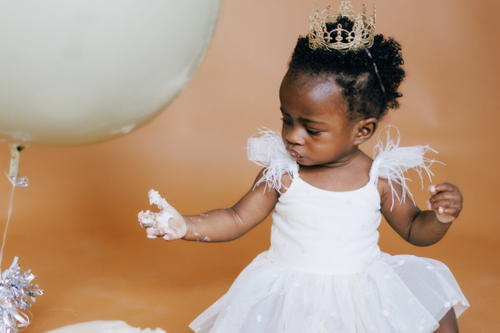 baby girl in white princess dress and crown with cake on her hand