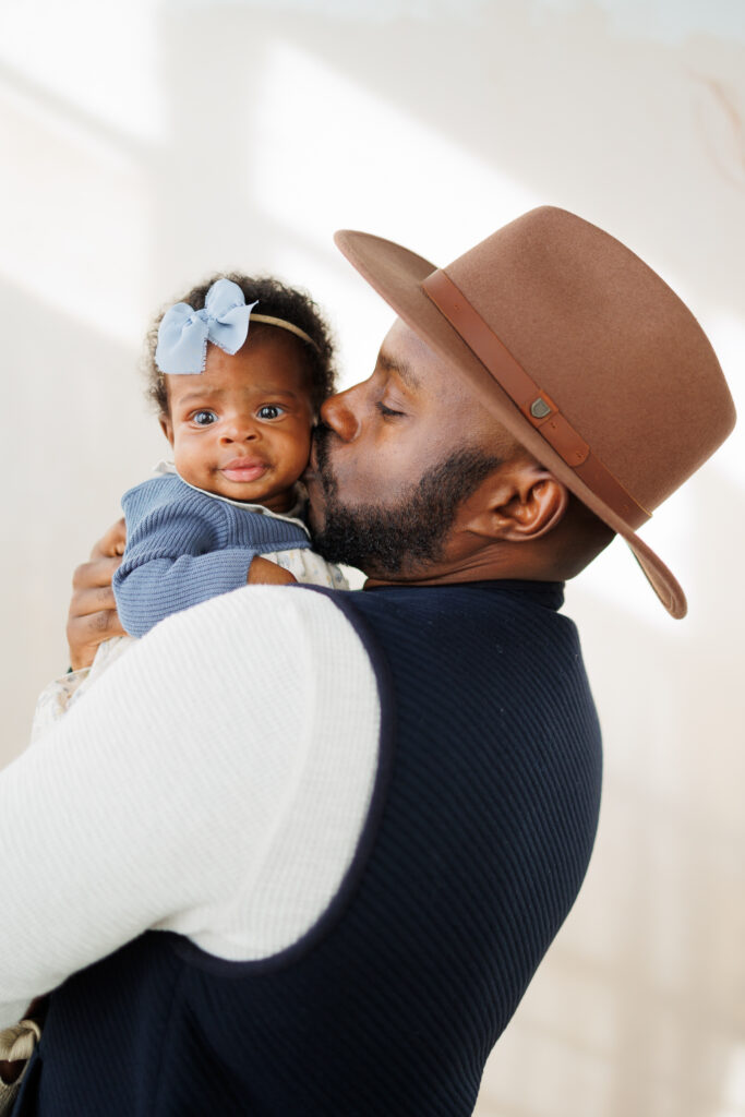 father holding three month old baby girl wearing blue bow against cream sunny backdrop. Father wearing brown felt hat and kissing newborns cheek