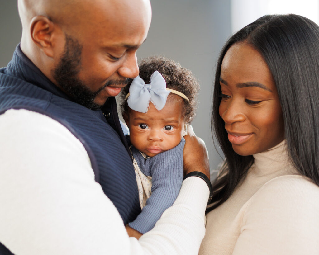 mother and father holding three month old baby girl and looking at her baby wearing blue bow and dress