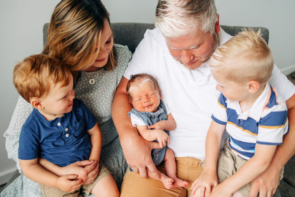 family with three sons and mother and father look at youngest newborn son while wearing shades of blue on a gray couch. Columbus pediatric Dentist