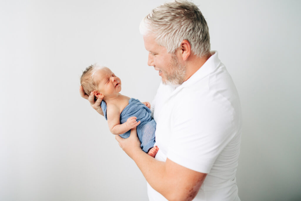 father and newborn baby son with matching white hair birthmarks look at each other as father holds son against a white backdrop