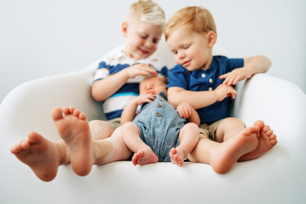 Two big brothers who are five and three hold newborn baby brother in white chair while thier feet are facing the camera. Columbus pediatric dentist