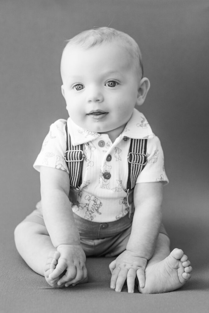 six month old little boy wearing collared shirt and suspenders sitting up holding feet looking at the camera in black and white photo