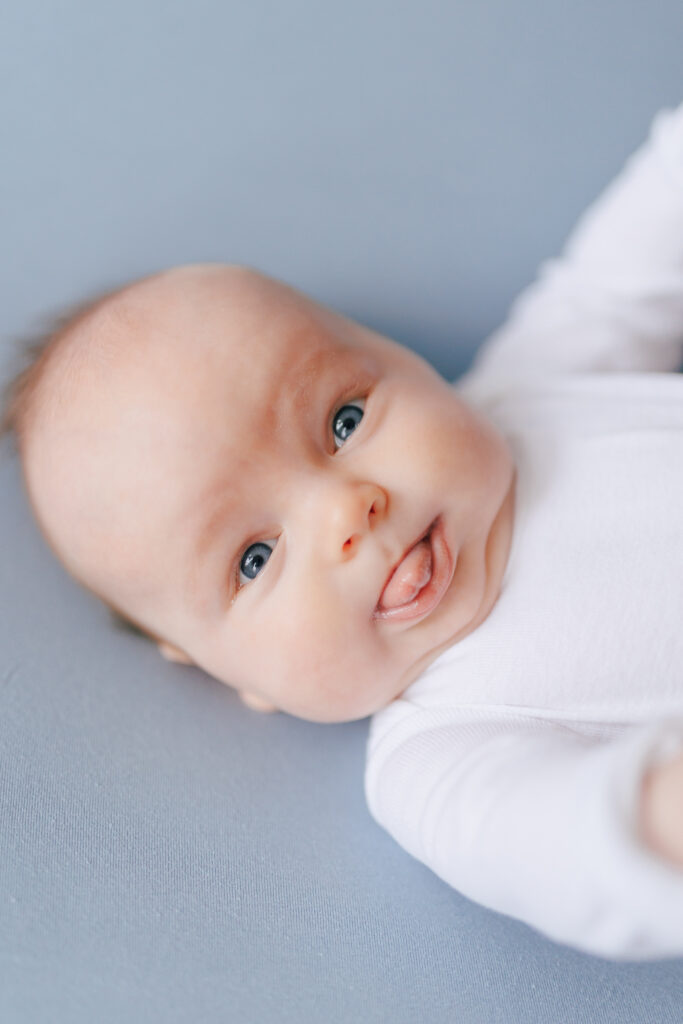 three month old baby boy on blue backdrop with blue eyes and white onsie sticking out his tongue looking at the camera