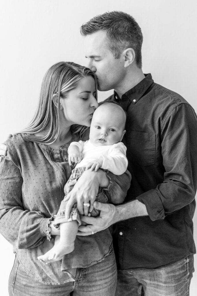 mother and father holding three month old son looking at photo in black and white photograph father is kissing mothers head and mother is kissing babies head