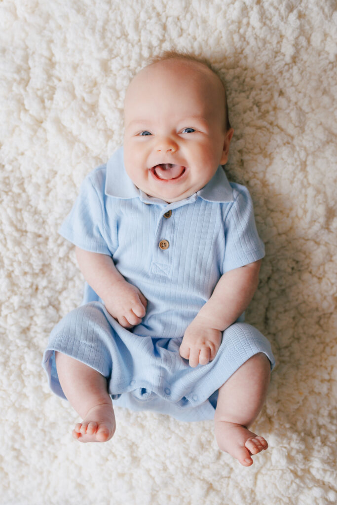 three month old boy in baby blue romper laying on back with huge smile at camera on cream fur backdrop