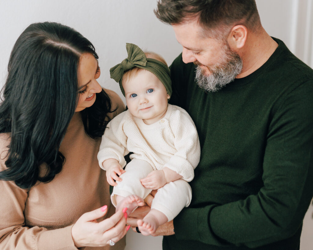 mother and father hold six month old daughter and smile at her while she smiles at the camera against white backdrop