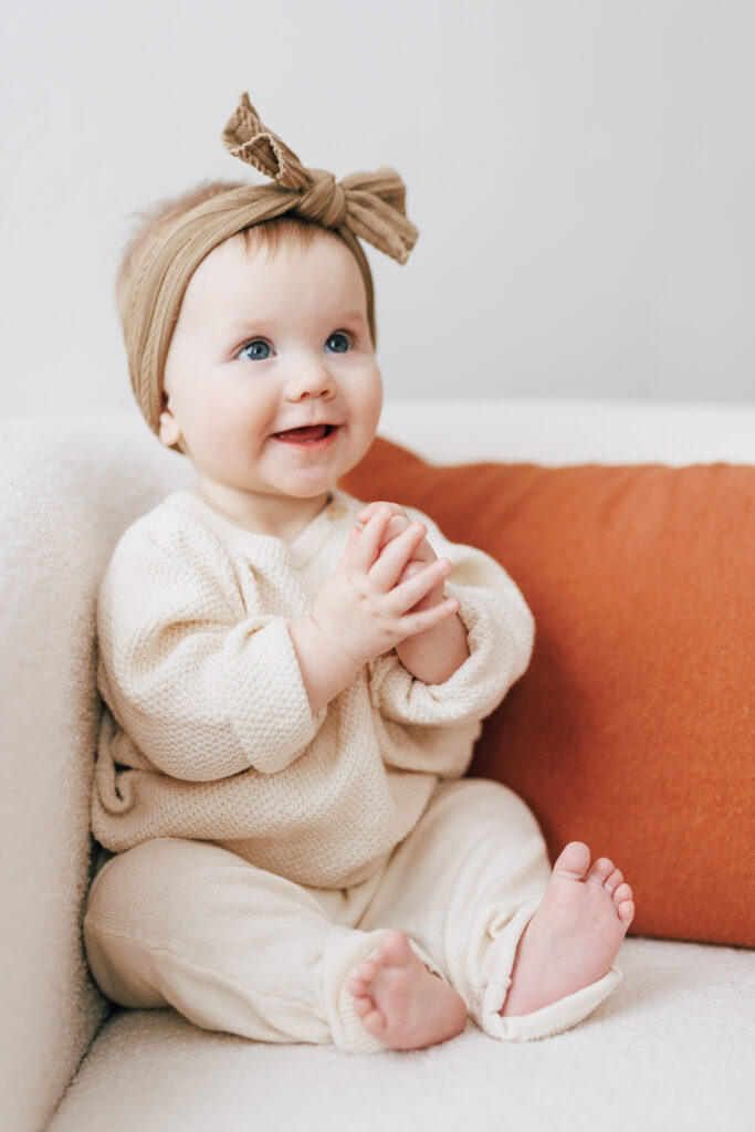 six month old girl sitting in white chair with rust colored pillow clapping in barefeet looking for columbus daycares