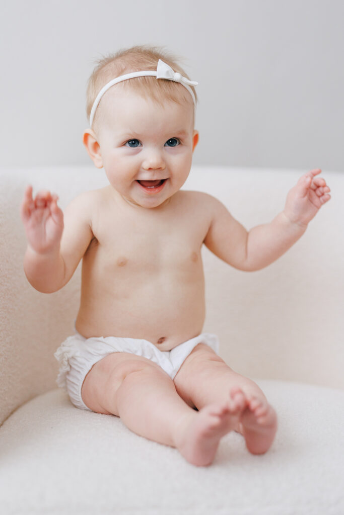 Little girl sitting in white chair six months old smiling in white diaper and white hairbow