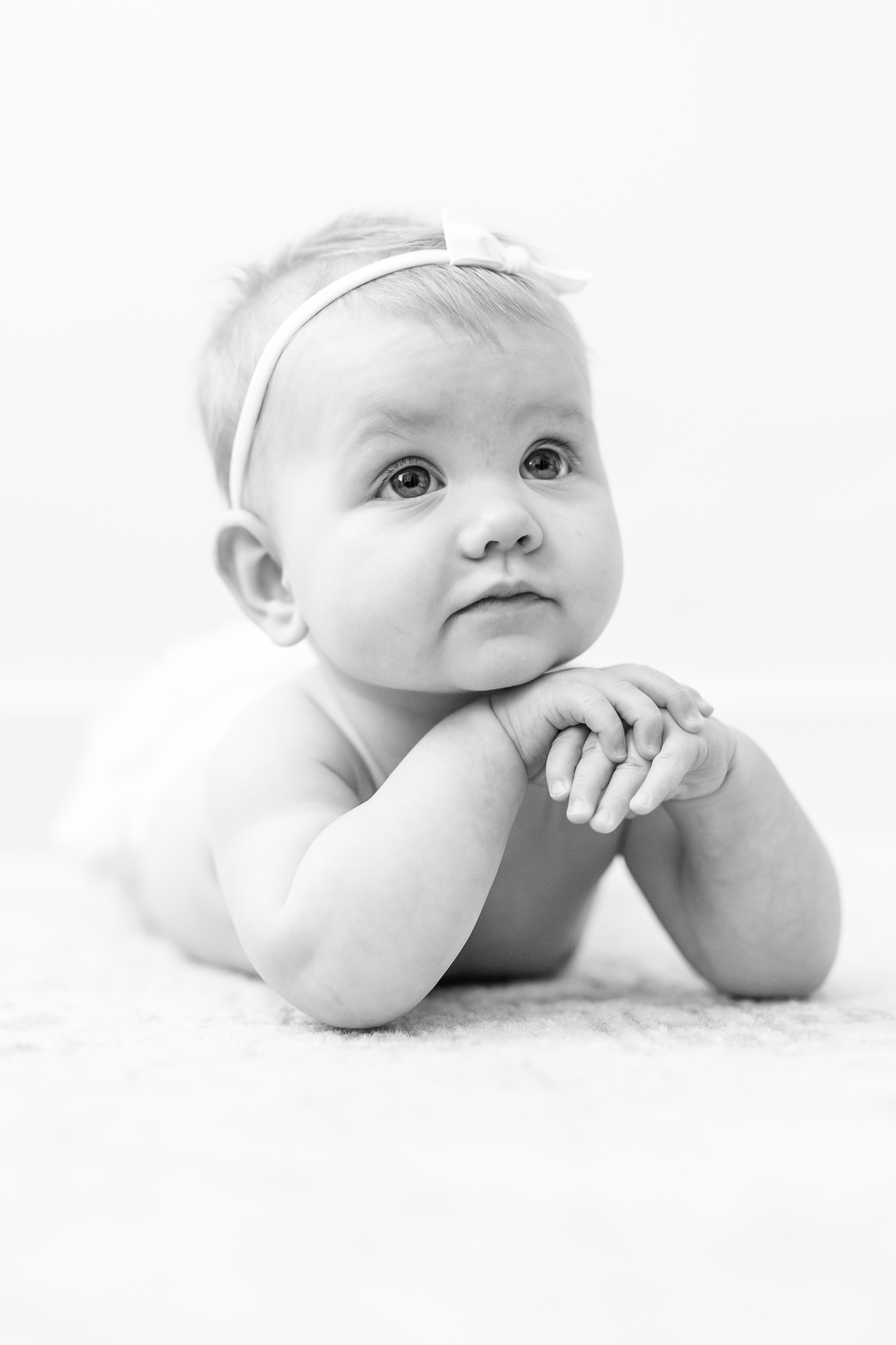 six month old little girl photograph in black and white with baby wearing white bow laying on her tummy with her hands folded under her chin thinking about parenting classes in Columbus
