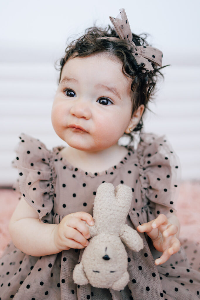 six month old wearing polka dot taupe dress and sitting up holding a teddy bear